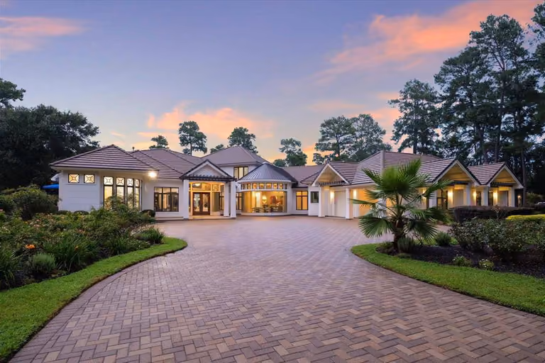 Twilight at a Memorial Villages estate home: warm light in the windows, a circular paver motor court in the foreground, and tall pines rising behind the roofline.